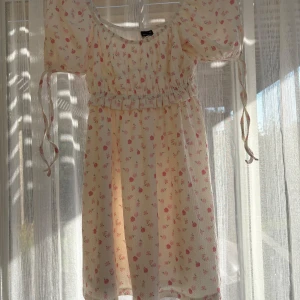 Blommig kortklänning från Gina Tricot - Supersöt kortklänning från Gina Tricot i ljus cremefärg med små rosa blommor. Puffärmar med snörning och smock i midjan ger en romantisk vibe. Klänningen har rund hals och är gjord i ett lätt, luftigt material som passar perfekt till varma dagar.