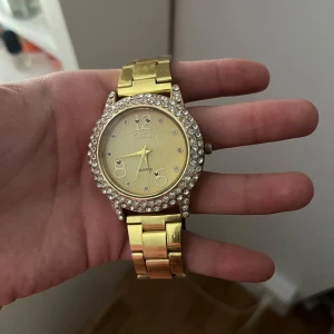 Guldig klocka med stenar från Kanima - Säljer en snygg guldig klocka från Kanima med glittrande stenar runt urtavlan. Klockan har en metallänk i guldton och en stilren urtavla med dekorativa siffror. Perfekt accessoar för dig som gillar bling och vill sticka ut.