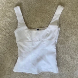 Vit korsett-topp  - Snygg vit korsett-topp  breda axelband och fyrkantig urringning. Figurnära passform som framhäver midjan och sömmar framtill för en stilren look. Perfekt att styla med jeans eller kjol för en trendig outfit.