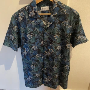 Blommig skjorta från Lindbergh - Snygg kortärmad skjorta från Lindbergh med tropiskt blommönster i blått och grönt. Skjortan har klassisk krage och knäppning framtill. Perfekt för dig som vill sticka ut med en färgstark och cool look.