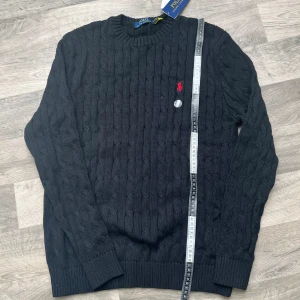 Svart kabelstickad tröja Polo Ralph Lauren - Kabelstickad svart tröja från Polo Ralph Lauren med klassisk röd broderad logga på bröstet. Tröjan har rund halsringning, ribbade muddar och är långärmad. Tillverkad i mjuk bomull, perfekt för lager-på-lager under kyliga dagar.