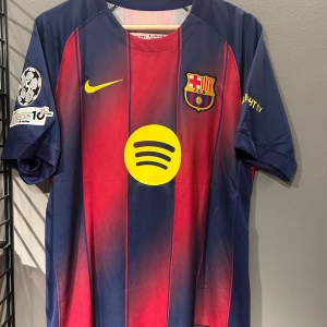 FC Barcelona Lamine Yamal #10 tröja - Säljer en FC Barcelona matchtröja med Lamine Yamal och nummer 10 på ryggen. Tröjan är randig i mörkblått och rött med gula detaljer, inklusive Spotify-logga och klubbmärke på bröstet. Kortärmad och tillverkad i lätt, ventilerande material. Perfekt för dig som älskar Barca! Helt ny med tagg en replika