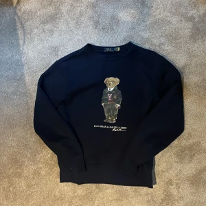 Mörkblå sweatshirt Polo Ralph Lauren - Snygg mörkblå sweatshirt från Polo Ralph Lauren i storlek M med ikoniska Polo Bear-trycket på bröstet. Tröjan har rund hals och långa ärmar, perfekt för dig som gillar klassisk streetstyle med en twist. Materialet är mjuk bomull som känns skönt mot huden. Pris ej hugget i sten