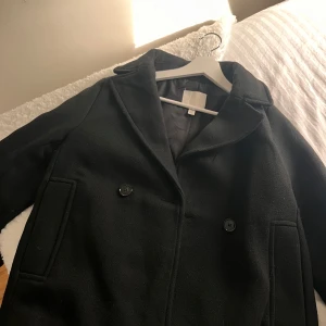 Svart rock från H&M XS - Klassisk svart kappa från H&M i storlek XS. Kappan har snygg krage, två stora fickor och stängs med knappar framtill. Köptes för 699kr säljer för 250 har bara använts 4 gånger men modellen är för lång för mig därför säljer jag den (jag är 1.55)