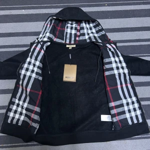 Svart Burberry hoodie med rutig luva - Helt ny Svart hoodiejacka från Burberry med dragkedja och klassiskt rutig insida i huva och foder. Jackan har två fickor fram, ribbade muddar och snörning i huvan. Tillverkad i 100% bomull och har den ikoniska Burberry-looken. Perfekt för dig som vill ha en stilren streetwear-vibe.