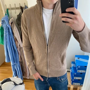 Beige kofta med helzip Dressmann - Säljer en beige kofta från Dressmann i storlek M. Koftan har dragkedja, ribbade muddar och hög krage. Tillverkad i mjukt stickat material, perfekt för lager-på-lager och chillare dagar. Snygg och enkel att matcha med jeans eller chinos.