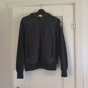 Svart fleece- och pufferjacka Parajumpers - Svart jacka från Parajumpers i kombinerat fleece- och puffer-material. Ordinarie pris 4000 kr. Jackan har huva, dragkedjor framtill och på fickor samt en snygg patch på ärmen. Perfekt för dig som vill ha en sportig och stilren look med extra värme. Den har en liten skada på framsidan som knappt syns hör av dig ifall du vill kolla närmre på den. 
