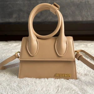 Beige handväska från Jacquemus - Superfin beige handväska från Jacquemus med rundade handtag och axelrem. Väskan har en stilren design med guldigt Jacquemus-logo framtill och är gjord i slätt skinn. Perfekt för dig som gillar trendiga accessoarer med unik form.