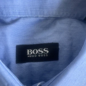 Blå skjorta från Hugo Boss, strl 42 - Snygg ljusblå skjorta från Hugo Boss i klassisk modell med krage, bröstficka och vita knappar. Skjortan är långärmad och tillverkad i mjuk bomull, perfekt för en clean och stilren look.