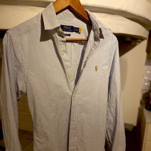 Randig skjorta från Polo Ralph Lauren - Säljer en klassisk slim fit skjorta från Polo Ralph Lauren i vitt och blått med tunna ränder. Skjortan har lång ärm, knappar framtill och den ikoniska broderade loggan på bröstet. Perfekt för dig som gillar stilrena och tidlösa plagg.