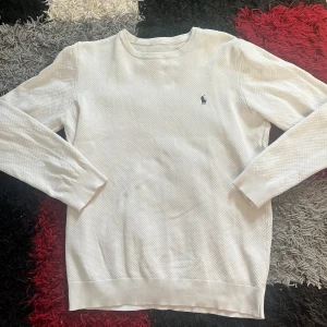 Vit stickad tröja från Ralph Lauren - Vit stickad tröja från Ralph Lauren med diskret strukturmönster och klassisk rund hals. Tröjan har lång ärm och den ikoniska broderade loggan i mörkblått på bröstet. Perfekt för dig som gillar stilren och enkel design.