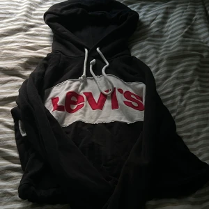 Svart Levi’s hoodie  - Svart hoodie från Levi's i storlek XS med stor vit och röd broderad logga över bröstet. Tröjan har huva med vita snören och är långärmad. Den är ganska kort så man visar magen men den ska sitta så💕