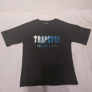 Svart Trapstar t-shirt med blått tryck - Svart t-shirt från Trapstar i storlek S /M med stort broderat Trapstar-tryck i vitt och ljusblått och mörkblått på bröstet. Klassisk passform med korta ärmar och rund hals. Materialet är mjuk bomull som känns skön mot huden. Perfekt för dig som gillar streetwear och vill sticka ut.