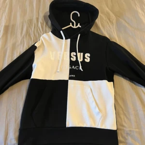 Svartvit hoodie från Versus Versace - Säljer en snygg hoodie från Versus Versace i svart och vitt blockmönster. Tröjan har huva med dragsko, stor magficka och tryckt logga på bröstet. Perfekt för dig som gillar streetstyle och vill sticka ut lite extra.