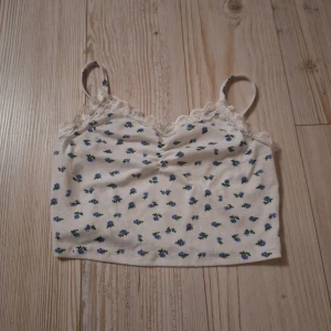 Vit blommig linnetopp från Shein - Supersöt kort vit linnetopp från Shein med små blå blommor och gröna blad över hela tyget. Toppen har spetskant vid halsen, smala axelband och en lätt rynkad detalj framtill. Materialet är ribbad bomull som känns mjukt mot huden. Perfekt för en chill sommarlook!