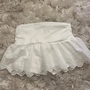 Vit kortkjol med spetsvolang - Supersöt vit lågmidjad kortkjol med bred linning och volangkant i broderad spets. Kjolen har ett romantiskt och fräscht uttryck med fina detaljer i spetsen och blommönster. Perfekt för varma dagar och somriga outfits.