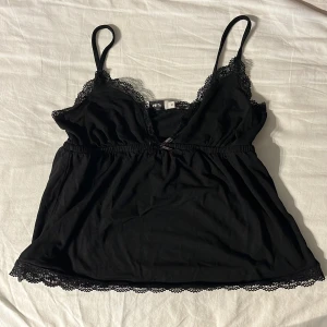 Svart spetslinnetopp från Nelly XS - Svart linnetopp från Nelly i storlek XS med tunna axelband och v-ringning. Toppen har fina spetsdetaljer längs urringning och nederkant samt en liten rosett framtill. Perfekt att styla lager på lager eller till jeans.