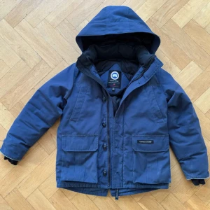 Blå jacka från Canada Goose - Säljer en blå dunjacka från Canada Goose med huva och stora fickor framtill. Jackan har klassisk patch på ärmen och stängs med både dragkedja och knappar. Perfekt för kalla dagar och har en riktigt clean look.