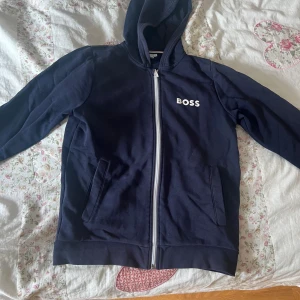 Marinblå hoodie från Hugo boss - Snygg marinblå hoodie från BOSS med vit logga på bröstet och hel dragkedja framtill. Tröjan har huva, två fickor och är långärmad. Skick 6/10 säljer den billigt. Storlek xs/12yrs. Mitt pris 100kr, vid fler frågor eller intresse skriv!