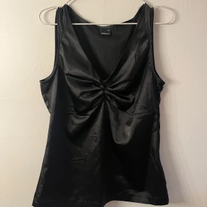 Svart satinlinnetopp  - Snygg svart linnetopp i satin från Gina Tricot med v-ringning och twistad detalj framtill. Ärmlös modell med glansig finish som ger en lyxig känsla. Perfekt att styla med jeans eller kjol för en trendig look.