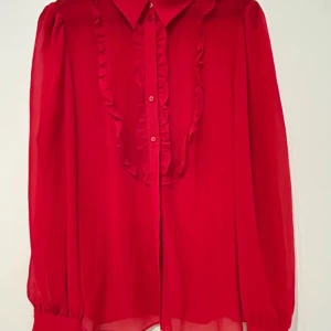 Röd volangskjorta från LIMÉ - Snygg röd skjorta från LIMÉ med volangdetaljer längs knappraden och klassisk krage. Skjortan har långa ärmar med breda manschetter och är tillverkad i ett lätt, luftigt material som ger en flowy känsla.