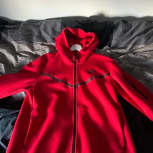 Röd Nike tech fleece - Röd Nike tech fleece i storlek M.. Nike vindjacka i rött med svart logga på bröstet och svarta detaljer. Jackan har hel dragkedja, huva och en ficka med dragkedja på ärmen. Perfekt för dig som vill ha en sportig och snygg look. Priset kan diskuteras vid snabb affär, inga defekter, knappt använd.