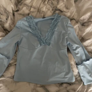 Ljusblå långärmad topp med spets - Supersöt ljusblå topp med långa ärmar och djup v-ringning dekorerad med spets. Snygg passform och mjukt material som känns skönt mot huden. Perfekt för dig som gillar romantiska detaljer och en feminin look.