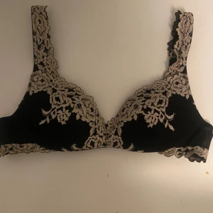 Svart spets-bh med blommigt mönster - Snygg svart bh med beige spetsdetaljer och blommigt mönster. Axelbanden är dekorerade med spets och bh:n har hakknäppning bak. Perfekt för dig som gillar en feminin och elegant stil. 
