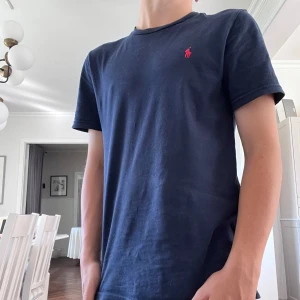 Mörkblå t-shirt från Polo Ralph Lauren - Snygg mörkblå t-shirt från Polo Ralph Lauren med klassisk röd logobrodering på bröstet. Modellen är normal passform med rund hals och korta ärmar. Tillverkad i mjuk bomull som är skön mot huden. Perfekt för dig som gillar stilrena och tidlösa plagg.