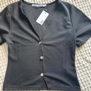Svart kortärmad blus från lager 157 - Svart blus med korta ärmar och snygg v-ringning från lager 157. Toppen har tre silvriga knappar framtill och är figurnära i modellen. Perfekt att styla med jeans eller kjol för en clean look. Pris går att diskutera.