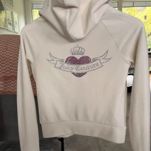 Vit hoodie från Juicy Couture!! - Säljer en vit hoodie från Juicy Couture med glittrande rosa hjärta och krona på ryggen, samt ett litet hjärta framtill. Tröjan har huva, snörning och dragkedja, samt två fickor fram. 