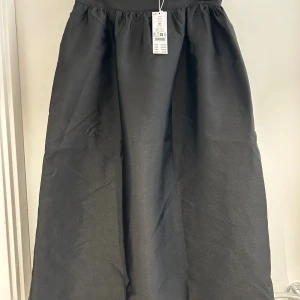 Svart långkjol från Gina Tricot - Säljer en svart långkjol från Gina Tricot med klassisk, vid passform och lätt glansigt tyg. Kjolen har en enkel och stilren look med rynkad midja som ger en snygg siluett. Perfekt att styla med sneakers eller boots.