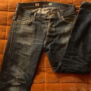 Nudie Jeans blå denimjeans - Snygga blå jeans från Nudie Jeans med klassisk femficksdesign och kontrastsömmar. Jeansen har en rak passform och tydliga slitningar för en cool, använd look. Bakfickorna har Nudies ikoniska broderi och läderpatch i midjan. Pris kan diskuteras.