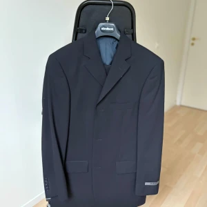 Strellson kostym – mörkblå (stl 148) - Elegant mörkblå kostym från kvalitetsmärket Strellson. Tillverkad i en lyxig ullblandning med stretch för bekväm passform. Klassisk design som fungerar lika bra till arbete som till fest och högtidliga tillfällen.  Storlek: 148 (passar lång & smal)  Färg: Mörkblå (nästan svart)  Modell: Novara-Luigi  Material: 93% virgin wool, 7% nylon  Nypris: 3995 kr  Skick: Helt ny med tags kvar
