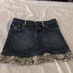 Jeanskjol med volang från KappAhl - Mörkblå jeanskjol från KappAhl med snygga slitningar, fickor fram och bak samt cool volangkant i grått och vitt camouflagemönster. Kjolen har bälteshällor och dekorativa detaljer på bakfickorna. Perfekt för dig som gillar en edgy och lekfull stil. Den går inte att köpa i butik längre.