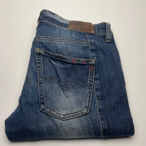 Blå jeans Replay Radula W27 L30 - Snygga blå jeans från Replay, modell Radula. Klassisk femficksdesign med slitningar och kontrastsömmar. Jeansen har röda detaljer vid fickorna och är tillverkade i bomull med en skön jeanskänsla. Perfekta för en avslappnad och cool stil.