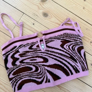 Rosa och brun mönstrad topp Bershka - Säljer en snygg croppad topp från Bershka i rosa och brunt med swirl-mönster. Toppen har smala axelband, en liten cut-out med ringdetalj framtill och är stickad i mjukt material. 