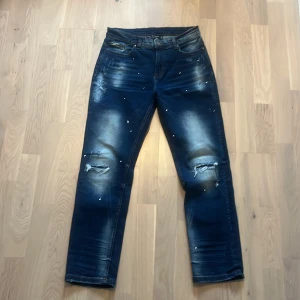 Blå jeans med slitningar Supply & Demand - Snygga blå jeans från Supply & Demand med slitna detaljer och hål på knäna. Jeansen har en cool tvätt med ljusare partier och små dekorativa nitar. Klassisk femficksmodell och normal passform, perfekta för en trendig streetstil.
