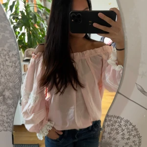 Ljusrosa offshoulder blus med spets - Supersöt ljusrosa blus med offshoulder-modell och vida ärmar. Blusen har fina spetsdetaljer på ärmarna och volangkant upptill. Perfekt för dig som gillar romantisk och luftig stil. Passar till både jeans och kjol.