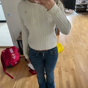 Vit kabelstickad tröja från GANT - Kabelstickad vit tröja från GANT med rund hals och diskret logga på bröstet. Tröjan har långa ärmar och ribbade muddar. Perfekt för lager-på-lager och passar till både jeans och kjol.