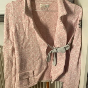 Odd Molly rosa cardigan jacka med knyt - Superfin rosa och vit cardigan jacka från Odd Molly med diskret mönster, lång ärm och snygg knytning framtill. Broderad detalj på ärmen med texten 'Lost on Purpose' och dekorativa fickor. Perfekt för dig som gillar en söt och avslappnad stil.