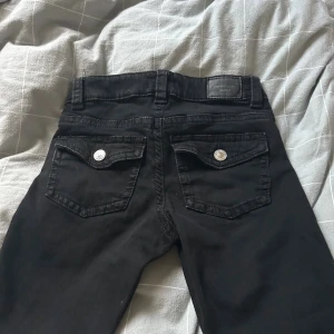 Svarta bootcut jeans från Perfect Jeans - Svarta jeans från Perfect Jeans i storlek 36. Modellen har bootcut-ben, klassiska fem fickor och snygga bakfickor med lock och silverknappar. Jeansen är i stretchigt bomullsmaterial och har normal passform med lowwaist