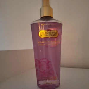 Victoria's Secret Strawberries & Champagne - En söt och fruktig body mist från Victoria's Secret med doft av jordgubbar och champagne. Flaskan är genomskinlig med rosa vätska och guldiga detaljer på etiketten och spraymunstycket. Perfekt för dig som gillar fräscha och bubbliga dofter.