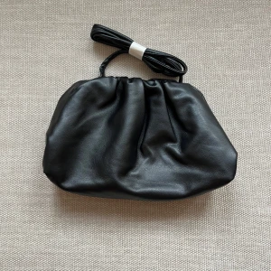 Dumpling bag - Stilren svart axelväska med mjuk, rynkad design och avtagbar axelrem. Väskan har en klassisk form och stängs med metallbåge upptill. Perfekt för dig som gillar minimalistisk och trendig look.