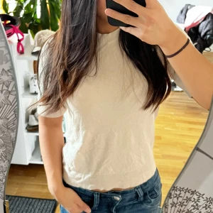 Beige stickad kortärmad topp - Säljer en enkel och stilren beige stickad topp med korta ärmar och rund hals. Toppen har en mjuk och lätt ribbad kant nertill och vid ärmsluten. Perfekt att matcha med jeans för en clean look.