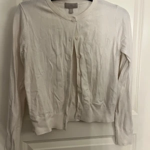 Vit kofta från H&M med knappar - En klassisk vit kofta från H&M med rund halsringning och knäppning framtill. Långärmad modell i mjukt och tunt material.