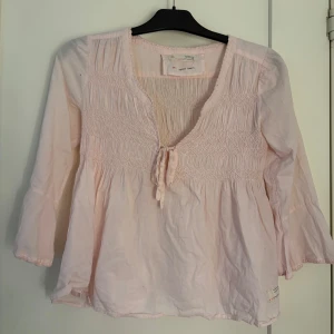 Ljusrosa Odd Molly blus med knyt - Supersöt ljusrosa blus från Odd Molly med v-ringning och knyt framtill. Vida ärmar med spetskant.