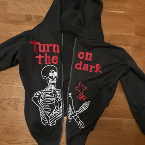 Svart hoodie med skelett och tryck - Svart hoodie med huva och dragkedja framtill. Framsidan har ett stort vitt skelett-tryck och röda bokstäver med texten 'turn the dark on'. Hoodien har långa ärmar och ribbade muddar. Perfekt för dig som gillar edgy och unik streetstyle.
