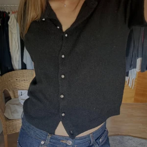 Svart stickad blus med pärlknappar - Säljer en svart stickad blus med korta ärmar och snygga pärlknappar framtill. Modellen är croppad och har en klassisk krage. Perfekt att styla med jeans för en chill men ändå uppklädd look.
