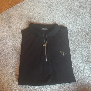 Svart pikétröja från Prada med zip - Svart pikétröja från Prada med klassisk krage och halv dragkedja framtill. Tröjan har korta ärmar och diskret rutstruktur i tyget samt Pradas ikoniska metallogo på bröstet. Perfekt för dig som gillar stilrena och exklusiva plagg.